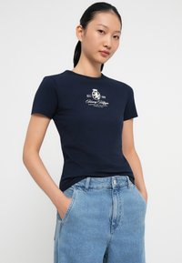 Tricou din bumbac navy cu mânecă scurtă, având un logo alb pe piept. Purtați-l cu blugi din denim de un albastru deschis, cu talie înaltă.