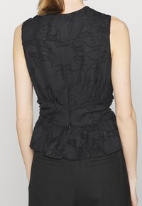 Musta ilmanhiha toppi, jossa on teksturoitu kukkakuvio, koottu vyötärö ja hillitty peplum-tyyli. Selässä on pieni vetoketjukiinnitys.