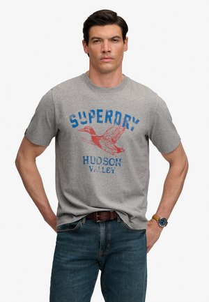 Mężczyzna w szarej koszulce Superdry Hudson Valley, niebieskich dżinsach, brązowym pasku i niebieskim zegarku, stojący z rękami w kieszeniach.