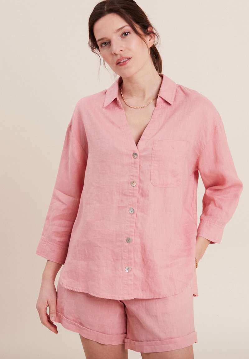 Camicia button-up in lino rosa chiaro con taschino sul petto e maniche a tre quarti, abbinata a pantaloni corti in lino coordinati, con orli arrotolati.