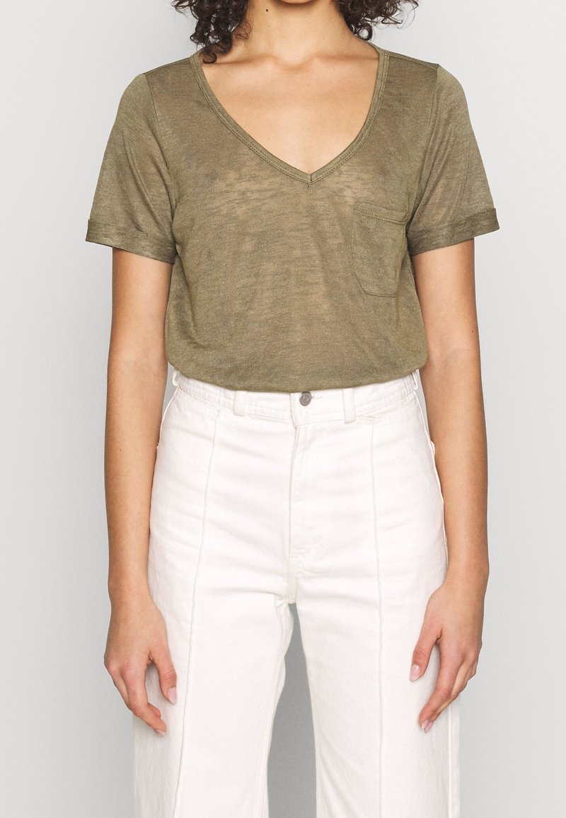 Femme portant un t-shirt vert olive à manches courtes avec col en V et poche poitrine, et un pantalon blanc taille haute sur un fond uni.