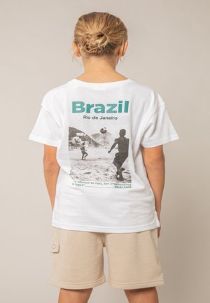 Enfant aux cheveux blonds réunis en chignon, portant un t-shirt blanc avec une scène de soccer de plage à Rio de Janeiro, Brésil, et un short beige.