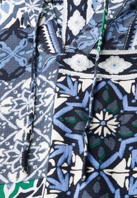 Blusa di cotone con un design in patchwork blu, nero e bianco con accenti verdi. Caratterizzata da intricati motivi geometrici e un cordoncino.