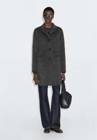 Grå uldfrakke med notch-krave, fire knapper og to frontlommer. Modellen bærer sorte solbriller og har en sort lædertaske.