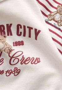 Hvidt bomuldsstof med bordeauxfarvet tekst og stribede detaljer. Indeholder en broderet teddybjørn og året "1989."