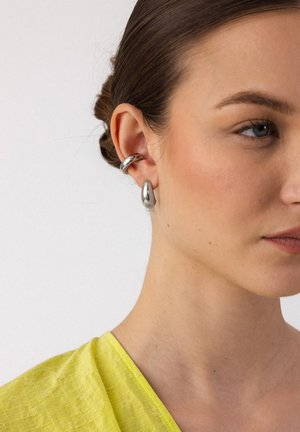 Junge Frau mit silbernen Creolen, die Haare zurückgebunden, gekleidet in einem gelben, strukturierten Oberteil vor einem schlichten Hintergrund.