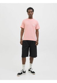 Lyserød t-shirt, korte ærmer med hvidt logo, parret med sorte knælange shorts. Bruges med sorte sneakers og hvide sokker.