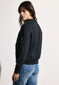 Svart bomberjacka med ribbat krage, blixtlåsficka på ärmen och elastiska muddar. Bärs över blå jeans, vilket visar en figurnära silhuett.