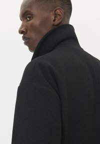 Čierny vlnený kabát s vysokým golierom, ktorého štruktúrovaný strih a hladká textúra sú zvýraznené čistým, minimalistickým dizajnom.