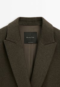 Gros plan sur le col et les revers d'un manteau en laine marron avec une étiquette intérieure indiquant Massimo Dutti, fabriqué au Portugal.