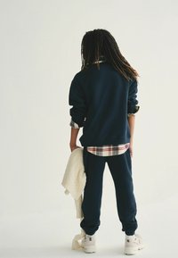 Sudadera de marina y pantalones de chándal, superpuestos sobre una camisa de cuadros. Suéter crema colgado sobre un brazo, zapatillas beige con suela texturizada.