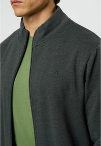 Chaqueta de cremallera gris oscuro con un alto cuello, que presenta un pequeño logo azul en el pecho, llevada sobre una camiseta verde. Tejido suave y texturizado.