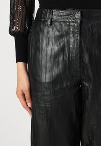 Sorte læderbukser med en tekstureret finish. Har en smal talje og sidelommer. Stoffet er glat med synlige folder og bølger.