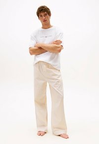 Jonge man met krullend haar staat blootvoets in een witte Tommy Hilfiger t-shirt en losse gele gestreepte broek tegen een effen achtergrond.