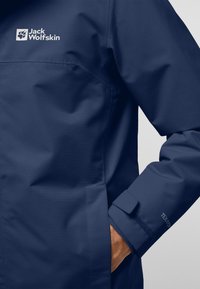 Giacca impermeabile blu navy con tessuto testurizzato, con logo sul petto e polsini regolabili. Mano parzialmente visibile nella tasca.