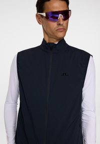 Mörkblå ärmlös väst med hög krage och dragkedja framtill, tillverkad av mjukt tyg, med en liten svart logotyp på bröstet och fickor med dragkedja.