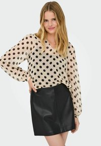 Gennemsigtig beige bluse med sorte prikker og lange ærmer, parret med en tætsiddende sort læder miniskirt. Glat tekstur og minimalistisk design.