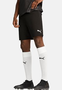 Schwarze Sportshorts aus atmungsaktivem Material mit einem weißen Puma-Logo. Kombiniert mit knielangen weißen Socken und schwarzen Fußballschuhen.