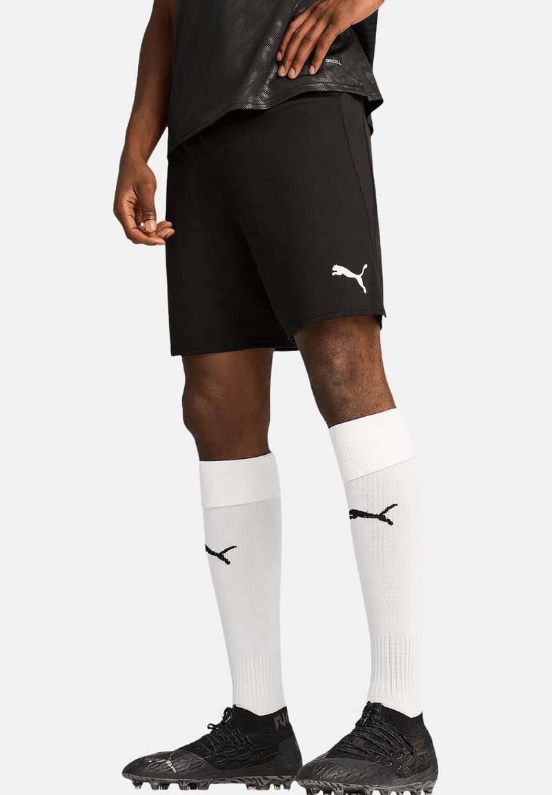 Schwarze Sportshorts aus atmungsaktivem Material mit einem weißen Puma-Logo. Kombiniert mit knielangen weißen Socken und schwarzen Fußballschuhen.