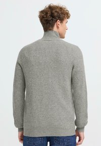 Pelēks svītrots džemperis ar augstu apkakli, teksturētu adījumu un garām piedurknēm. Mugurpusē ir taisna apmale, uzvilkts virs zilajiem džinsiem.