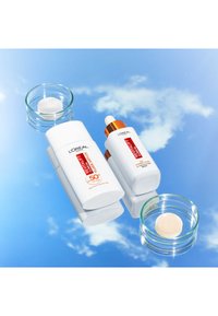 Produits de soins L'Oréal Revitalift : contenants blancs avec des accents orange, plat en verre rond avec de la crème, texture lisse, reflétant un fond de ciel bleu.