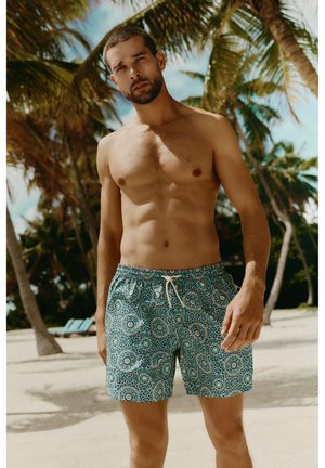 Skjorteløs mand iført mønstrede badeshorts står på en sandstrand med palmer i baggrunden under klar himmel.