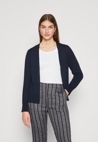 Cardigan bleu marine à ouverture devant, manches longues, poignets côtelés et deux poches avant, associé à une chemise blanche et un pantalon rayé.