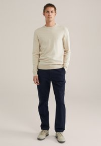 Uomo in piedi con le mani in tasca, che indossa un maglione beige, pantaloni blu navy e sneakers beige su uno sfondo semplice.