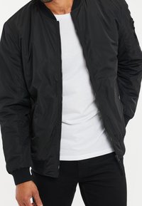 Giacca bomber nera con texture liscia, chiusura frontale con zip, tasche laterali, polsini a costine e vestibilità casual, indossata sopra una camicia bianca.