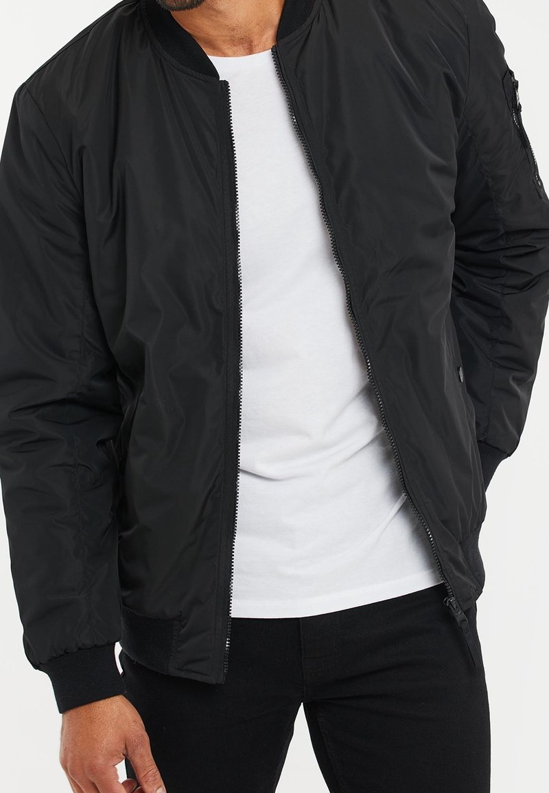 Giacca bomber nera con texture liscia, chiusura frontale con zip, tasche laterali, polsini a costine e vestibilità casual, indossata sopra una camicia bianca.