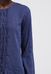 Mörkblå långärmad topp med texturerade vertikala veck längs framsidan, bärs av en person mot en enkel ljus bakgrund.