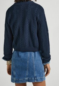 Navy knitted sweater with cable patterns, ribbed cuffs and hem, worn over a light denim skirt featuring visible stitching and pockets.