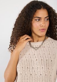 Top sans manches en tricot beige avec des lignes verticales et de petits trous, associé à un collier en chaîne argentée épaisse avec un anneau central.
