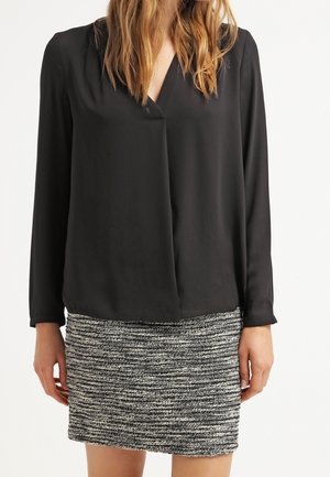 Vrouw die een zwarte blouse met lange mouwen en een V-hals draagt en een getextureerde grijs-witte tweed minirok.