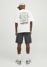 T-shirt en coton blanc avec un design graphique noir et le texte "originals studio" au dos, associé à un short cargo gris et une casquette beige.