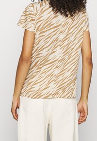 Beige kortärmad topp med ett brunt tigerstripe-mönster och rak skärning. Tyget verkar mjukt och har en enkel sömdetalj bak.