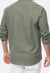 Camisa verde de manga larga con cuello abotonado y mangas remangadas, hecha de tela suave, con textura lisa y un corte relajado.