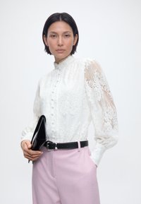 Γυναίκα που φοράει λευκή δαντελένια μπλούζα και ροζ παντελόνι, κρατώντας ένα μαύρο clutch, κοιτάζοντας μπροστά με αυτοπεποίθηση σε ένα απλό φόντο.