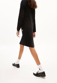 Robe noire mi-longue aux genoux avec manches longues, texture douce, fentes latérales et une étiquette en tissu près de l'ourlet. Associée à des mocassins noirs et des chaussettes blanches.