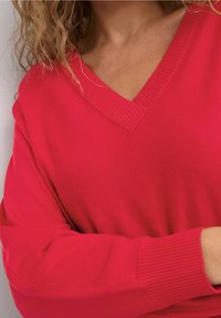 Frau trägt einen leuchtend roten V-Ausschnitt-Pullover mit gerippten Ärmelbündchen, zeigt die Schulter und hat die Arme verschränkt vor einem schlichten Hintergrund.