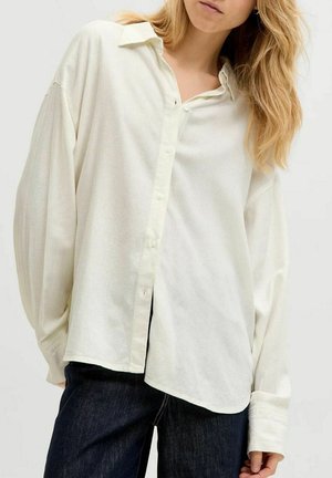 Mujer que lleva una camisa blanca de botones de ajuste holgado con mangas largas y vaqueros azul oscuro sobre un fondo liso.