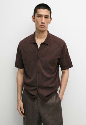 Uomo con capelli corti e neri che indossa una camicia marrone scuro a maniche corte con bottoni e pantaloni marroni a righe sottili su uno sfondo chiaro uniforme.