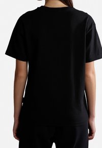 Persona vistiendo una camiseta negra de manga corta y pantalones negros, mostrada desde atrás sobre un fondo blanco.