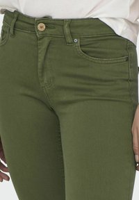 Jeans de mezclilla verde oliva con un diseño ajustado. Presentan un cierre delantero con botón, dos bolsillos frontales y costuras distintivas a lo largo de las costuras.