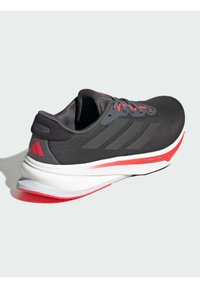 Black athletic shoe with a textured upper, gray accents, red laces, and a white midsole featuring a red stripe, designed for performance.