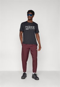 Fekete Nike póló logóval, bordó, tapered fazonú nadrág, fekete sportcipő és kék napszemüveg. Sima anyag, laza szabás és márkás részletek.