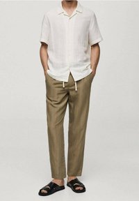 Bílá lněná košile s krátkým rukávem, béžové pruhované zužující se kalhoty a černé sandály. Texturovaná látka a ležérní střih. Nastavitelný detail v pase.