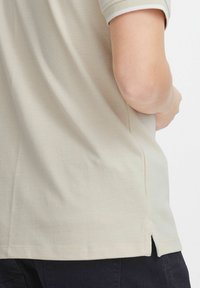 Beige polo skjorte med teksturert stoff, korte ermer med hvite mansjetter og sidesplitter for bedre bevegelsesfrihet.