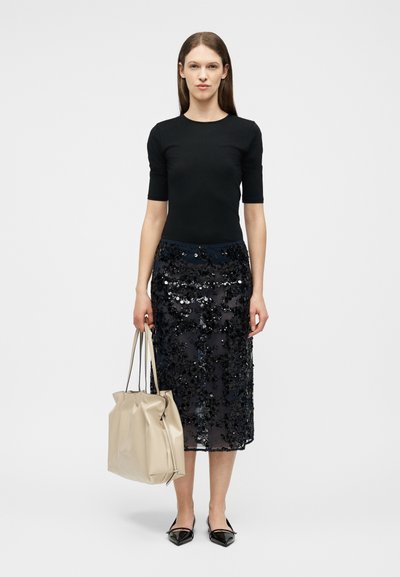 Femme en haut noir ajusté et jupe à sequins tenant un sac à main beige, debout devant un fond clair uni.