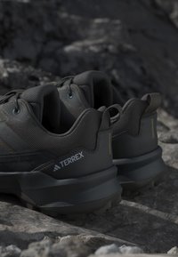 Chaussures de randonnée noires Adidas Terrex posées sur une surface rocheuse, montrant des collerettes rembourrées, des boucles et des semelles texturées pour une utilisation en extérieur.